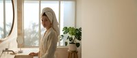 Young woman in bathrobe with towel on head standing in modern bathroom after shower. Morning self-care routine and wellness lifestyle concept.