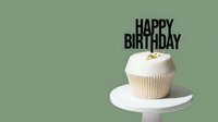 White frosted cupcake with a black Happy Birthday topper on a white stand, sage green background, minimalist style celebration dessert.