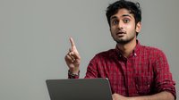 Young South Asian man in red plaid shirt gesturing with finger while working on laptop, expressing idea or solution against neutral background.