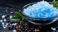 Blue bath salt crystals in glass bowl with fresh green leaves and scattered black peppercorns on dark textured background, spa and wellness concept.