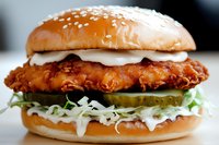 Crispy fried chicken sandwich with shredded lettuce, pickles, and creamy sauce on toasted sesame seed bun, close-up view showing layered ingredients.