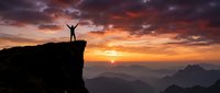 Silhouette of person with raised arms celebrating success on mountain cliff edge during dramatic sunrise with orange purple sky and layered peaks below.