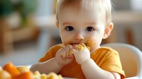 Caucasian baby in yellow t-shirt eating solid food in high chair at home. Close up portrait of infant during mealtime showing early childhood development and healthy nutrition habits.