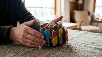 Senior man's protective hands surrounding diverse group of wooden figurines representing community care and social protection concepts.