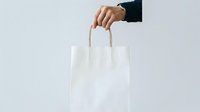 Male hand in dark business sleeve holding white paper shopping bag with handles against neutral background. Suitable for retail, commerce, shopping, and business marketing materials.