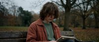 Young Caucasian teenage boy with messy hair reading a book on a park bench surrounded by autumn trees on an overcast day.