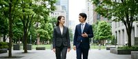 Asian business professionals, man and woman in suits, walking and talking outdoors in urban park setting with green trees and city buildings.