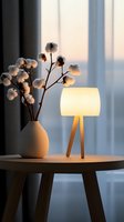 Cozy evening atmosphere with modern table lamp and cotton stems in ceramic vase on wooden side table against soft window light creating warm ambiance.