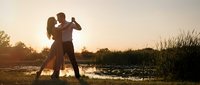 Romantic couple dancing together at golden hour sunset by lake with warm lighting creating intimate silhouette moment for wedding anniversary celebration.