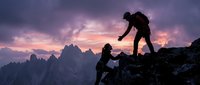 Silhouetted climber helping partner reach mountain summit at dramatic sunset with jagged peaks below, teamwork and adventure concept for outdoor sports and motivation.