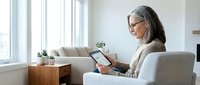 Mature woman with gray hair and glasses using tablet computer while sitting in modern armchair by window in bright living room interior.