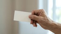 Senior adult male hand holding blank white card against neutral interior background, suitable for business messaging and promotional campaigns.