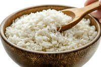 Steamed white rice served in wooden bowl with bamboo spoon, close up view showing fluffy texture and individual grains against white background.