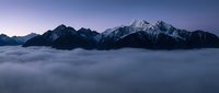 Majestic snow-capped mountain peaks rising above thick cloud layer during twilight hour creating dramatic alpine landscape with purple sky gradient.