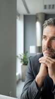 Thoughtful mature Caucasian businessman with gray hair and beard in dark suit contemplating in modern office environment with natural lighting.