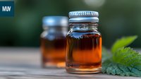 Glass jars filled with amber liquid and metal lids on wooden surface with green leaves and bokeh background, natural organic honey or maple syrup product photography.