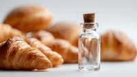Small glass bottle with cork stopper next to freshly baked croissants on white surface, perfect for food and beverage industry.