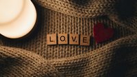 Wooden letter tiles spelling LOVE on a cozy knit sweater with a red heart and soft candlelight, warm romantic Valentine's Day mood.