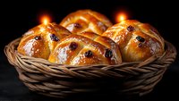 Fresh homemade sweet bread rolls with raisins in rustic woven basket, decorated with burning candles on dark background, traditional holiday baking.