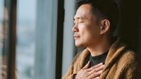 Asian man with eyes closed in peaceful meditation by window, wearing cozy brown cardigan, hand on chest in mindfulness practice or gratitude gesture.