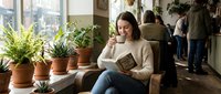 Young Asian woman reading book while sitting by window with potted plants in cozy cafe. Relaxed student enjoying literature in modern coffee shop interior.
