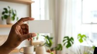 Person with dark skin tone holding blank white card in bright room with potted plants, ideal for business cards, invitations, and promotional designs.
