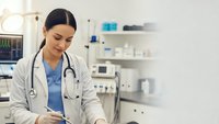 Young female doctor in white coat with stethoscope working on tablet in modern medical office. Healthcare professional using digital technology.