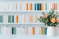 Minimalist bookshelf with color coordinated books in pastel tones and white ceramic vase with peach roses and greenery arrangement against white wall.