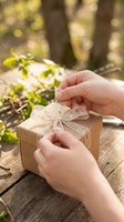 Child's hands unwrapping wooden gift box with translucent ribbon bow outdoors on wooden table surrounded by green foliage and natural sunlight, suitable for holiday, celebration and gifting concepts.