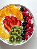 Fresh fruit platter with sliced oranges, kiwi, strawberries, raspberries and blackberries arranged on white cream in ceramic bowl on light background.