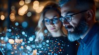 Caucasian couple illuminated by glowing blue digital particles, man with beard and glasses, woman with blonde hair, looking at magical light effects in dark room.