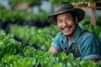 Asian male farmer in hat and overalls smiling while tending to organic lettuce crop in greenhouse. Natural lighting highlights sustainable agriculture practices.