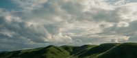 Dramatic rolling green hills beneath a moody sky filled with towering cumulus clouds, capturing the raw beauty of mountainous terrain for landscape and nature projects.