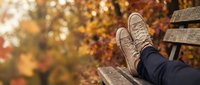 Person relaxing on wooden bench in autumn park with golden foliage, wearing casual sneakers and dark pants, peaceful outdoor lifestyle moment.