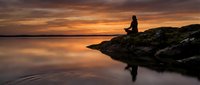 Silhouetted woman sitting on rocky outcrop at sunset, meditating by calm water with golden sky reflection and peaceful natural landscape.