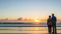Elderly couple standing together on beach watching golden sunset over ocean waves, romantic moment during vacation, peaceful evening by sea.