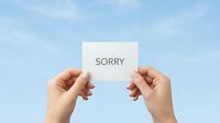 Hands holding a white card with the word Sorry against a blue sky, symbolizing apology, forgiveness, and reconciliation concepts.
