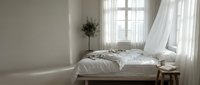 Minimalist bedroom interior with unmade bed, flowing white curtains and natural sunlight creating peaceful morning atmosphere for home decor and lifestyle concepts.