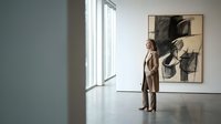 Woman in brown coat viewing abstract charcoal artwork in minimalist gallery space with natural light from large windows, suitable for art, culture, and interior design projects.