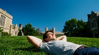 Young male student relaxing on campus lawn with hands behind head enjoying sunny day at university with historic buildings and clear blue sky.