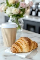 Fresh buttery croissant on white napkin with takeaway coffee cup on marble table, soft focus roses in glass vase creating cozy breakfast atmosphere.