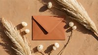 Elegant terracotta envelope surrounded by dried pampas grass and white flowers on textured beige background for wedding invitations and stationery.