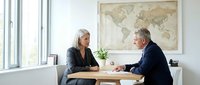 Professional business meeting between mature Caucasian woman and man discussing documents at modern office desk with world map background.
