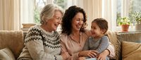 Three generations of women laughing together on a cozy sofa, grandmother, mother, and granddaughter sharing a joyful family moment at home.