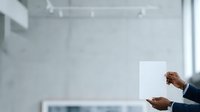 Person of African descent, male, wearing dark business attire, holding blank white paper or document in modern office environment with whiteboard and natural lighting.