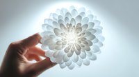 Delicate white paper flower with intricate layered petals held gently in hand against soft glowing light background for wellness and mindfulness concepts.