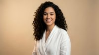 Young Hispanic woman with curly hair smiling confidently in white blazer against beige background for professional business portrait and corporate headshot.