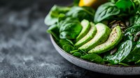Fresh spinach salad with sliced avocado and lemon on gray ceramic plate, dark background with copy space. Clean eating and healthy food concept.