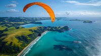 Aerial view of paraglider soaring over stunning coastal landscape with turquoise ocean waters, green rolling hills, and dramatic cliffs creating perfect adventure sports scenery.