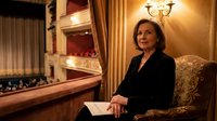 Elegant mature woman in black suit sitting in luxury opera house box with ornate golden interior and red velvet seats during evening performance.
